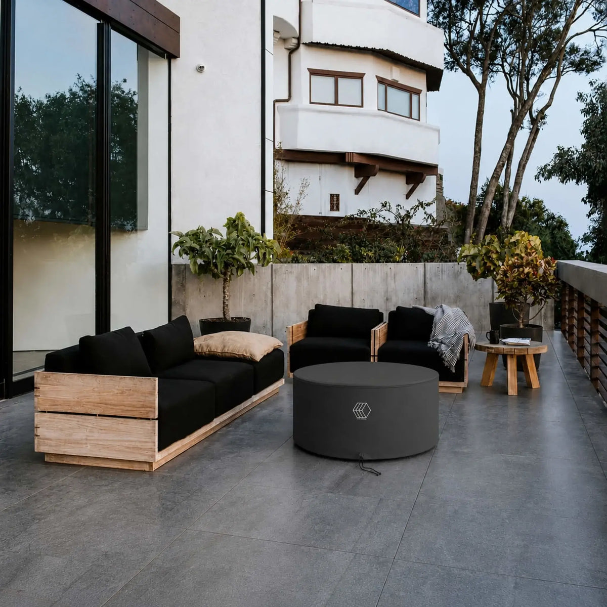 HPC Fire Hearth Products GFRC Concrete Belmont Weather Cover installed on a round outdoor fire pit table within a modern patio seating area. Surrounded by contemporary wood-framed furniture with black cushions, the black fitted weather cover protects the Belmont fire feature from exposure to the elements while maintaining a clean, sophisticated appearance. The cover blends seamlessly into the upscale outdoor living environment.