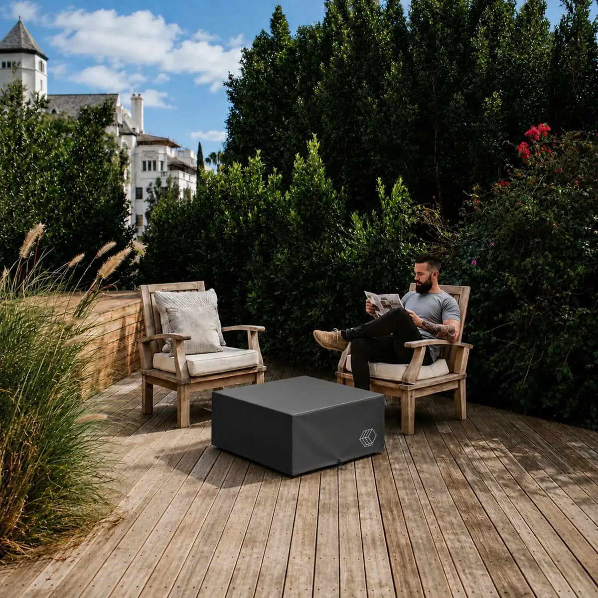 HPC Fire Hearth Products Controls GFRC Concrete Bravo 32 x 32 Weather Cover shown covering a square outdoor fire pit table on a wooden deck patio. Surrounded by comfortable lounge chairs, lush landscaping, and mature greenery, the black protective cover demonstrates how the Bravo 32 x 32 Weather Cover preserves fire pit surfaces and controls from rain, sun, and debris. The lifestyle setting showcases practical all-season protection within an inviting residential outdoor living environment.