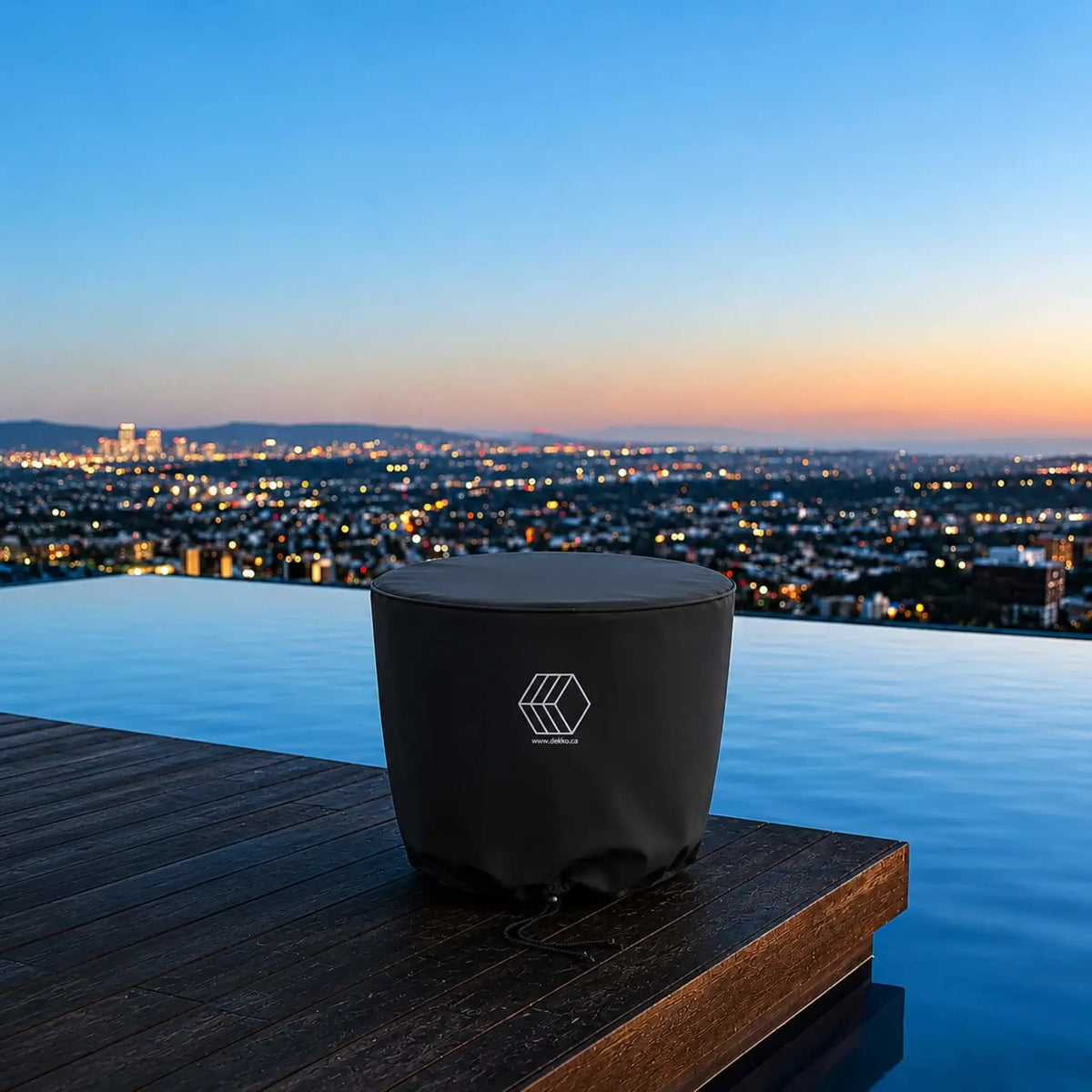 HPC Fire Hearth Products Controls GFRC Concrete Element 20 Round Fire Column Weather Cover Lifestyle 1 featuring a covered round fire column positioned on a luxury infinity-edge pool deck overlooking a panoramic city skyline at sunset. The black fitted weather cover protects the fire feature while blending seamlessly into the upscale outdoor living space. The image highlights year-round protection, premium outdoor design, and weather-resistant storage for modern fire column installations.