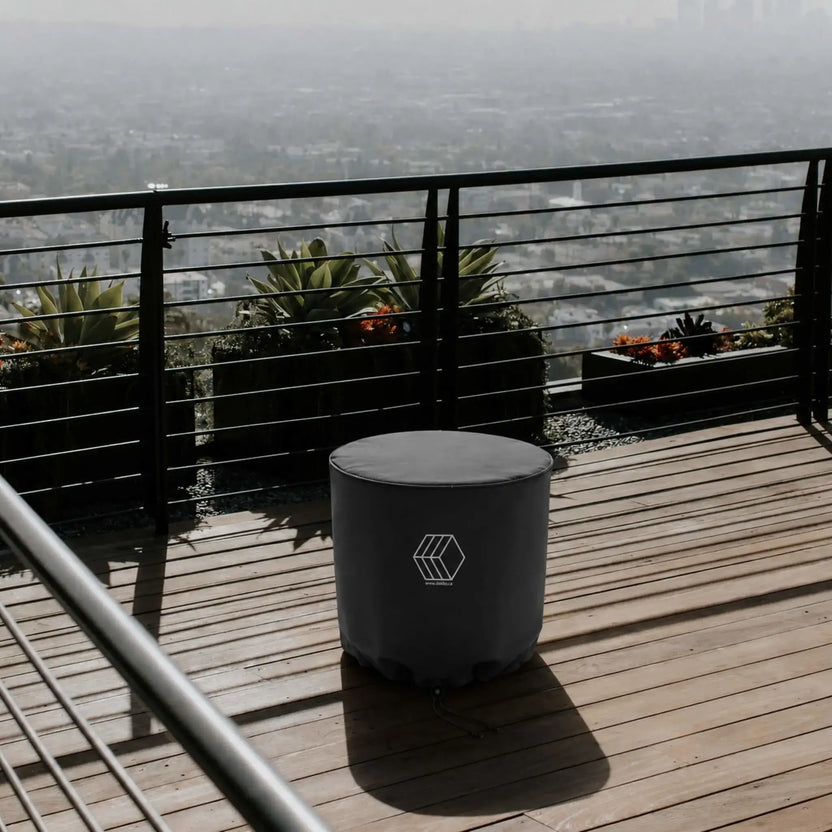 HPC Fire Hearth Products Controls GFRC Concrete Element 20 Round Fire Column Weather Cover Lifestyle 2 showing the protected fire column on a rooftop deck with wood plank flooring, metal railing, and elevated city views in the background. The custom-fitted black weather cover features Dekko branding and a secure drawstring bottom designed to shield the round fire column from outdoor elements. This lifestyle scene demonstrates practical protection while maintaining a clean, sophisticated patio aesthetic.