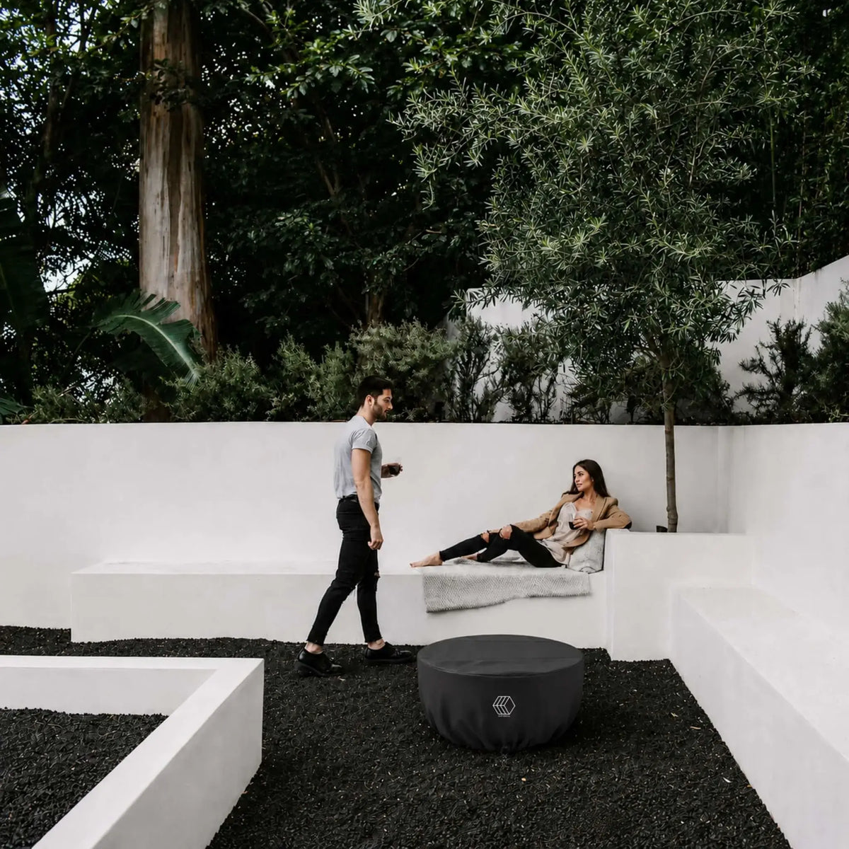HPC Fire Hearth Products Controls GFRC Concrete Serenade 32 Weather Cover Lifestyle 1 showing the covered fire pit in a modern outdoor courtyard. The black weather cover protects the Serenade fire pit while blending into a contemporary landscape featuring white architectural seating walls, dark decorative stone ground cover, mature trees, and outdoor lounge space. This lifestyle image highlights year-round protection and clean aesthetics for luxury residential environments.