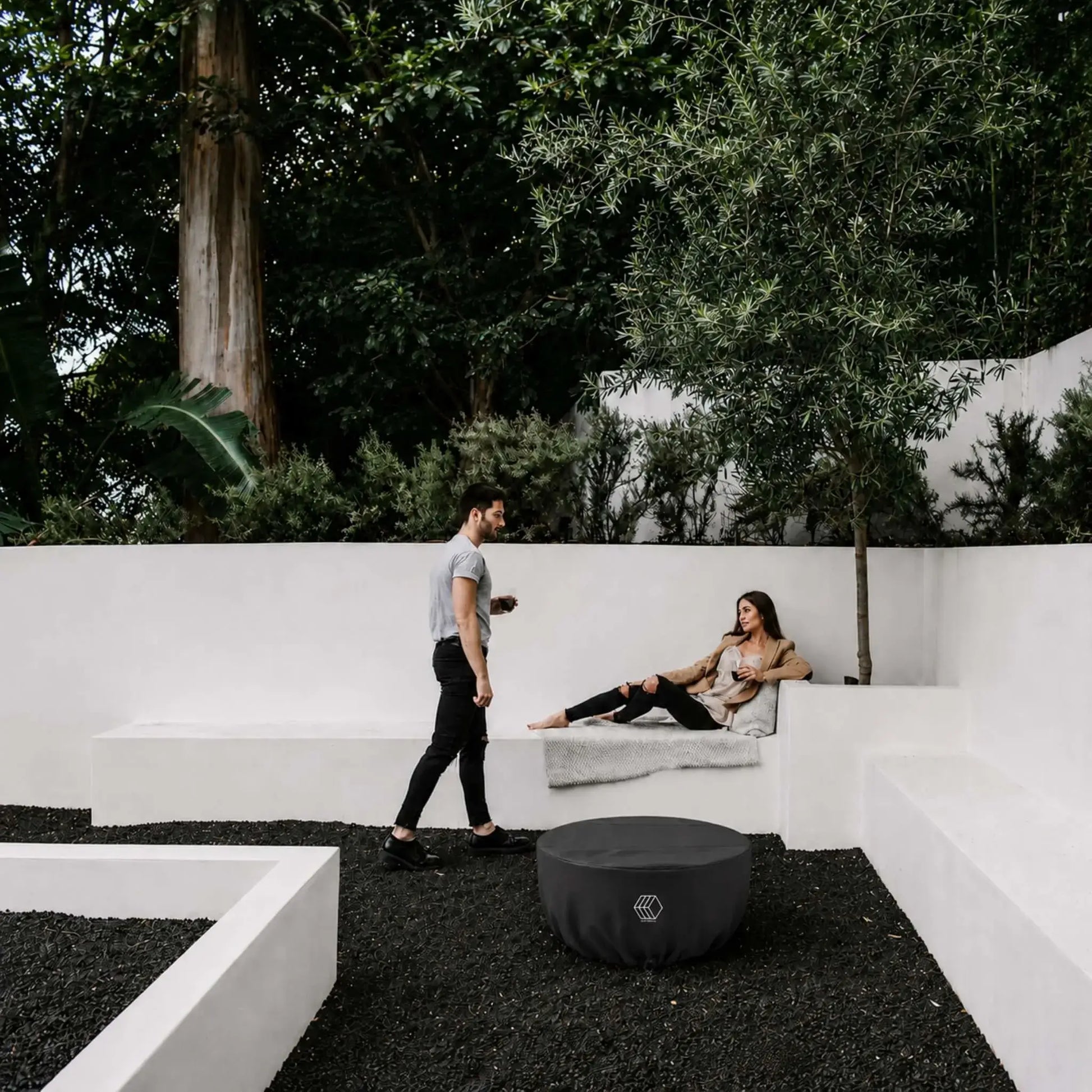 HPC Fire Hearth Products Controls GFRC Concrete Serenade 32 Weather Cover Lifestyle 1 showing the covered fire pit in a modern outdoor courtyard. The black weather cover protects the Serenade fire pit while blending into a contemporary landscape featuring white architectural seating walls, dark decorative stone ground cover, mature trees, and outdoor lounge space. This lifestyle image highlights year-round protection and clean aesthetics for luxury residential environments.