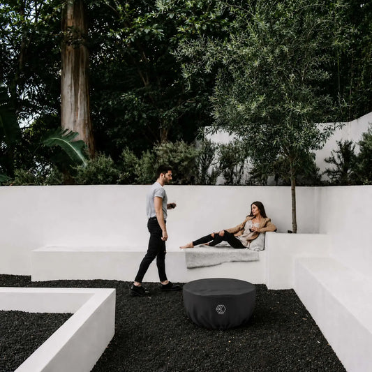 HPC Fire Hearth Products Controls GFRC Concrete Serenade 32 Weather Cover Lifestyle 1 showing the covered fire pit in a modern outdoor courtyard. The black weather cover protects the Serenade fire pit while blending into a contemporary landscape featuring white architectural seating walls, dark decorative stone ground cover, mature trees, and outdoor lounge space. This lifestyle image highlights year-round protection and clean aesthetics for luxury residential environments.