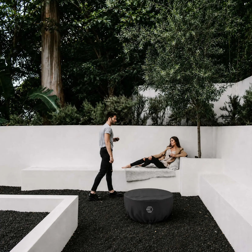 HPC Fire Hearth Products Controls GFRC Concrete Serenade 32 Weather Cover Lifestyle 1 showing the covered fire pit in a modern outdoor courtyard. The black weather cover protects the Serenade fire pit while blending into a contemporary landscape featuring white architectural seating walls, dark decorative stone ground cover, mature trees, and outdoor lounge space. This lifestyle image highlights year-round protection and clean aesthetics for luxury residential environments.