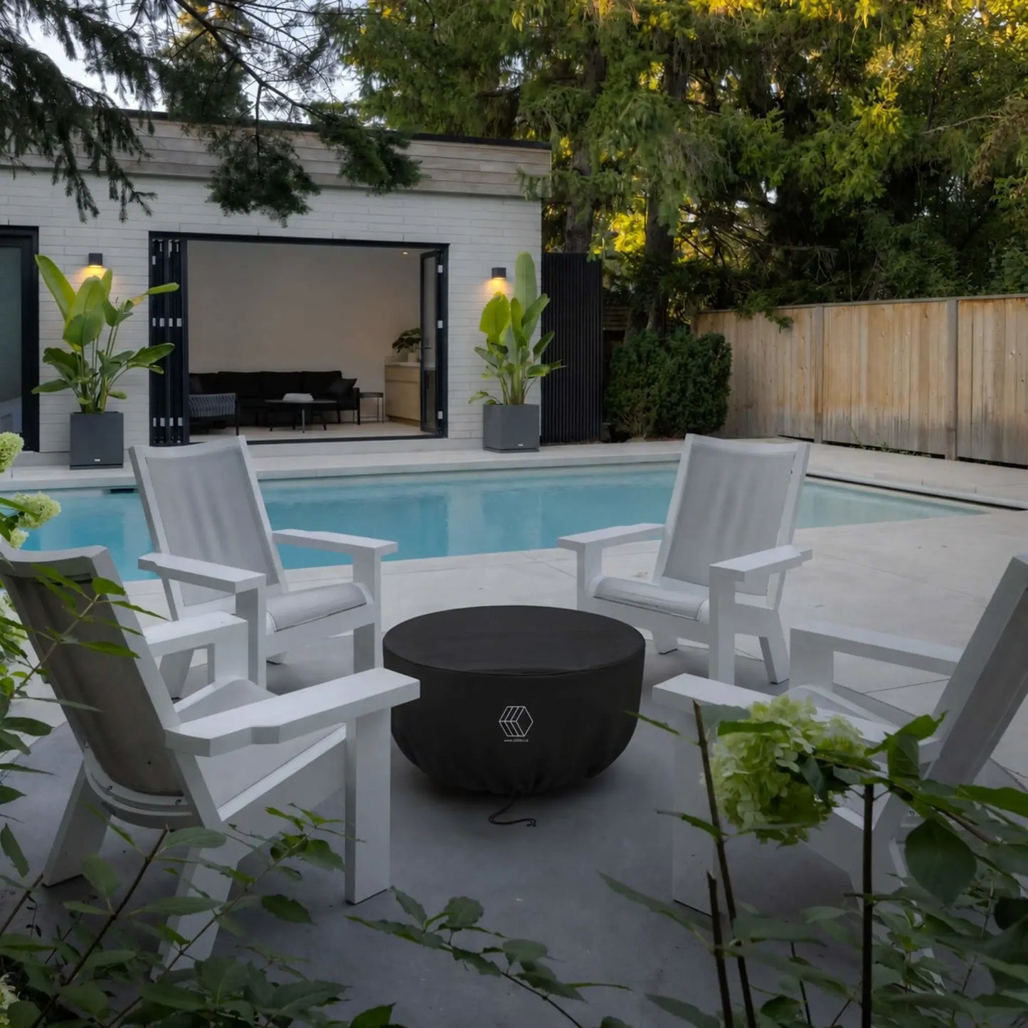 HPC Fire Hearth Products Controls GFRC Concrete Serenade 32 Weather Cover Lifestyle 2 featuring the covered round fire pit positioned between white Adirondack-style chairs beside a backyard swimming pool. The fitted black weather cover protects the Serenade fire pit from outdoor elements while maintaining a neat appearance in the seating area. Surrounded by landscaped greenery, a pool house, and modern patio furnishings, the image demonstrates practical fire pit protection in an upscale outdoor living space