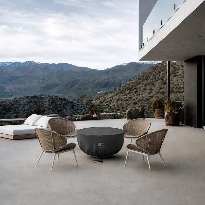 HPC Fire Hearth Products Controls GFRC Concrete Serenade 40 Weather Cover installed on a round outdoor fire pit within a luxury patio setting. The black fitted cover protects the Serenade 40 fire pit while surrounded by woven lounge chairs overlooking a dramatic mountain landscape. Designed for year-round outdoor protection, the weather cover helps guard the GFRC concrete fire feature from moisture, dirt, UV exposure, and changing weather conditions.