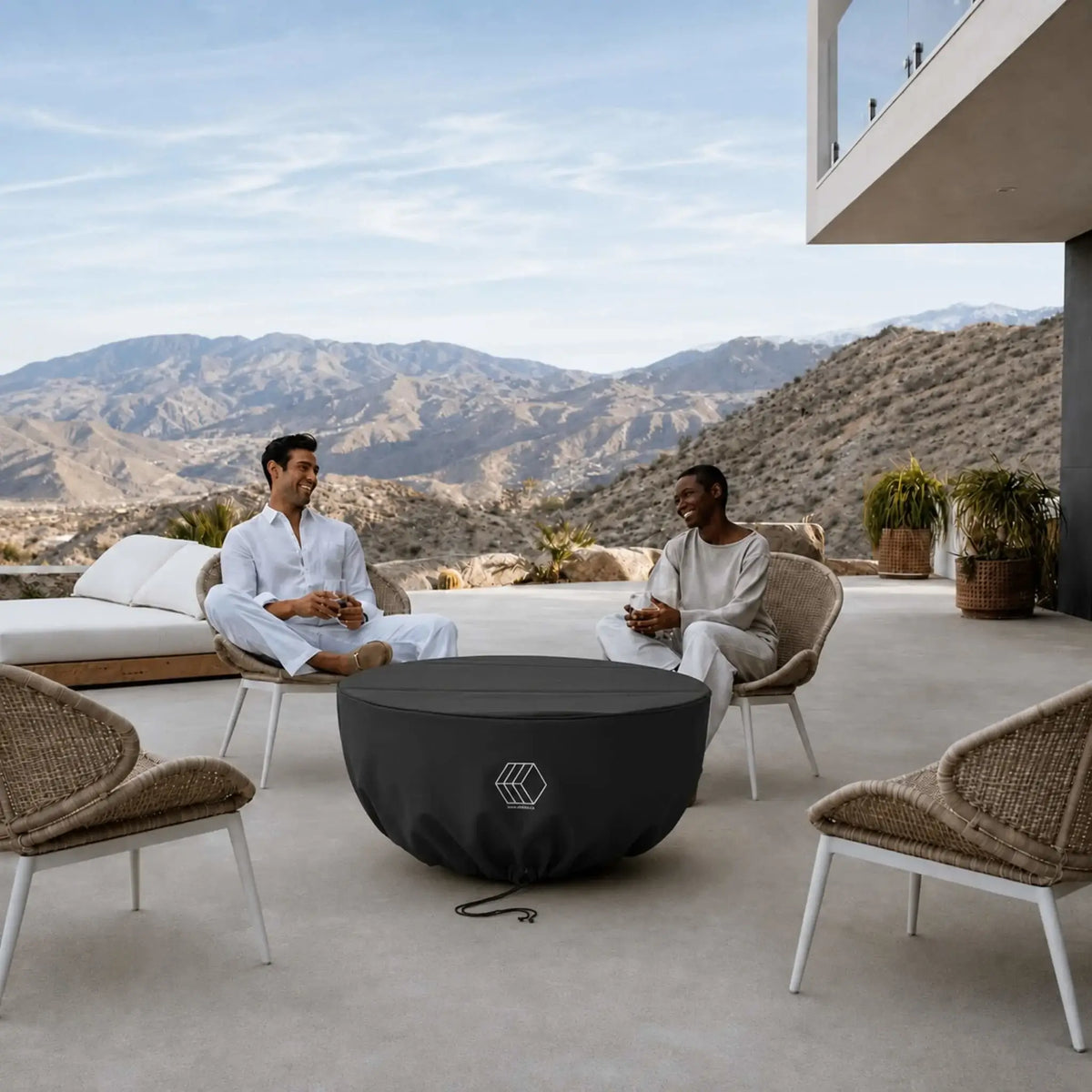 HPC Fire Hearth Products Controls GFRC Concrete Serenade 40 Weather Cover featured on a covered fire pit at a modern outdoor living space with panoramic mountain views. Two people relax in surrounding lounge chairs while the black weather cover securely protects the Serenade 40 fire pit between uses. The fitted cover includes a drawstring closure and durable construction, helping preserve the fire pit’s finish and internal components from outdoor exposure.