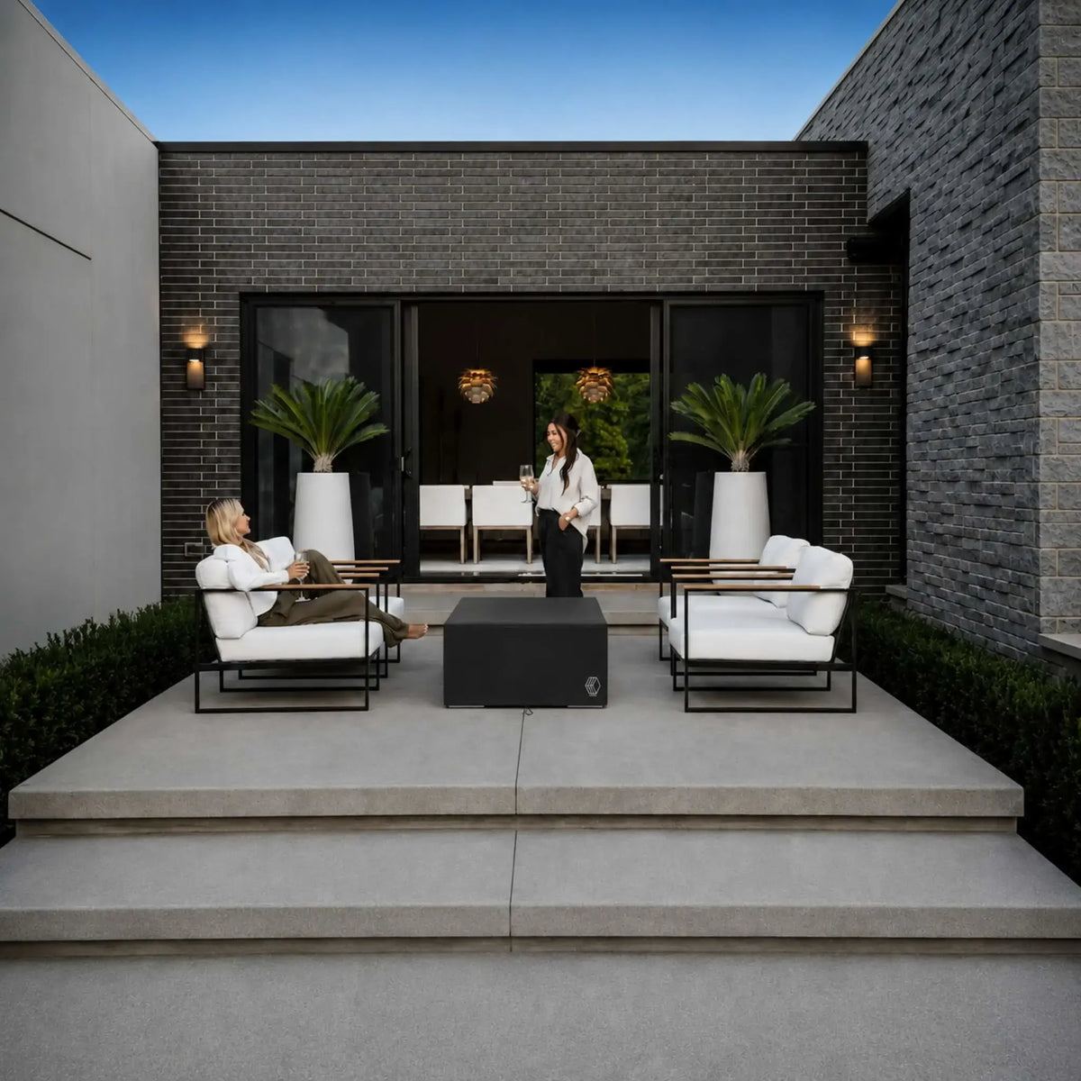 HPC Fire Hearth Products Controls GFRC Concrete Sonoma 56 x 30 Weather Cover Lifestyle 1 image showing the protective cover installed over a Sonoma fire pit in a modern outdoor living space. The black fitted cover sits between contemporary lounge chairs with white cushions on a raised patio. Two people relax and socialize nearby, demonstrating how the weather cover helps protect the fire feature while maintaining a clean, upscale patio aesthetic.