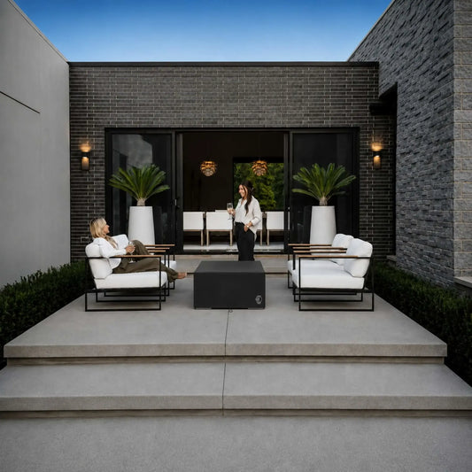 HPC Fire Hearth Products Controls GFRC Concrete Sonoma 56 x 30 Weather Cover Lifestyle 1 image showing the protective cover installed over a Sonoma fire pit in a modern outdoor living space. The black fitted cover sits between contemporary lounge chairs with white cushions on a raised patio. Two people relax and socialize nearby, demonstrating how the weather cover helps protect the fire feature while maintaining a clean, upscale patio aesthetic.
