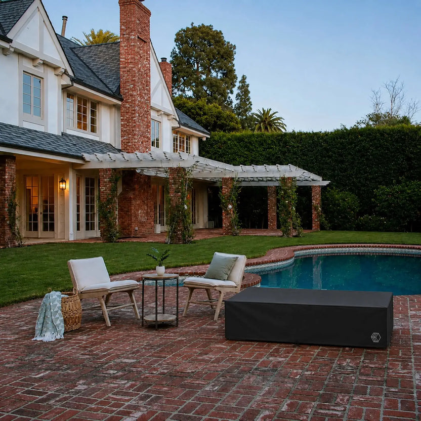 HPC Fire & Hearth Products Controls GFRC Concrete Sonoma 72 x 33 Weather Cover displayed on a covered fire pit table beside a swimming pool and lounge seating area. The black fitted weather cover protects the rectangular Sonoma fire pit from outdoor exposure while blending seamlessly into the upscale backyard setting. Surrounded by brick patio flooring, cushioned chairs, and a luxury home exterior, the cover demonstrates practical year-round protection for outdoor fire features.