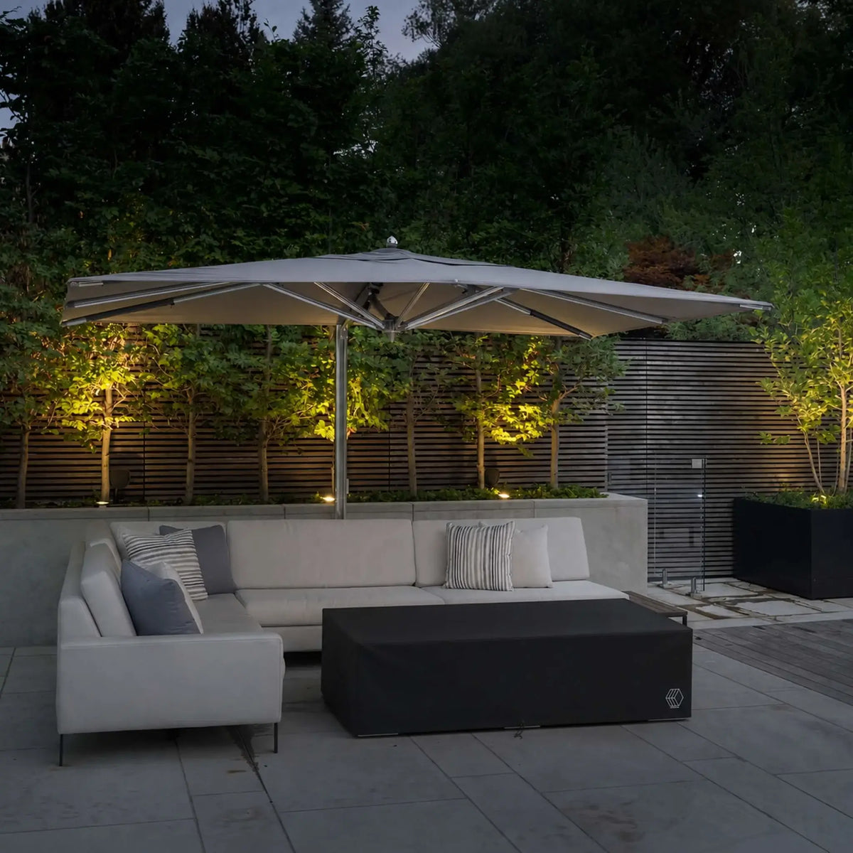 HPC Fire & Hearth Products Controls GFRC Concrete Sonoma 72 x 33 Weather Cover featured on a modern outdoor patio beneath a large cantilever umbrella. The black fitted cover fully encloses the Sonoma rectangular fire pit table, protecting it from moisture, dirt, leaves, and sun exposure when not in use. Surrounded by contemporary seating, landscape lighting, and architectural privacy screens, the cover complements the elegant outdoor living environment while providing dependable weather protection.