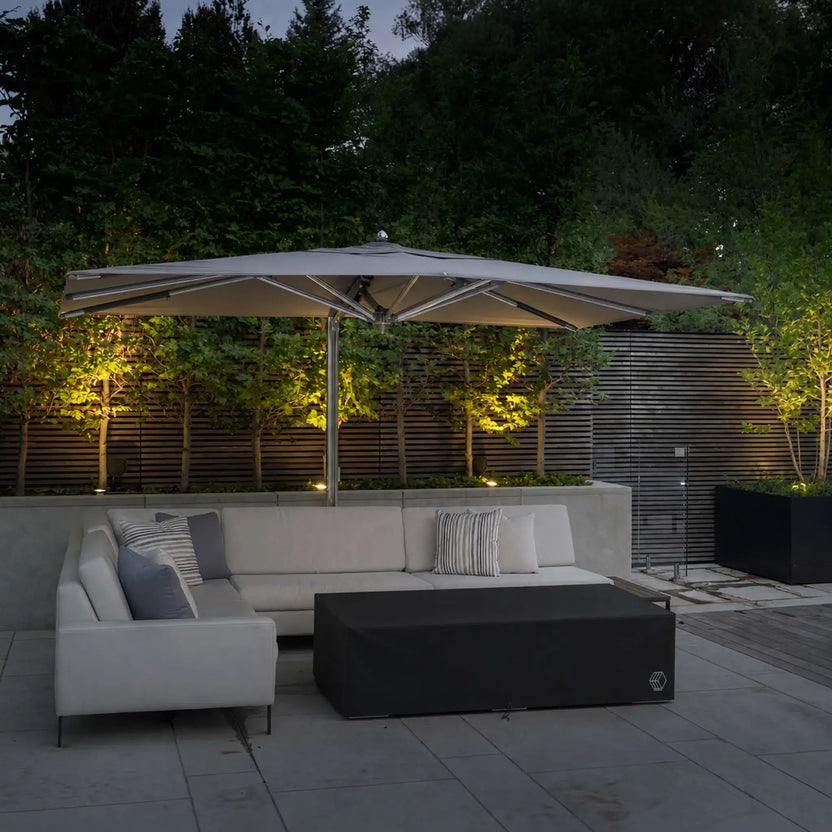 HPC Fire & Hearth Products Controls GFRC Concrete Sonoma 72 x 33 Weather Cover featured on a modern outdoor patio beneath a large cantilever umbrella. The black fitted cover fully encloses the Sonoma rectangular fire pit table, protecting it from moisture, dirt, leaves, and sun exposure when not in use. Surrounded by contemporary seating, landscape lighting, and architectural privacy screens, the cover complements the elegant outdoor living environment while providing dependable weather protection.
