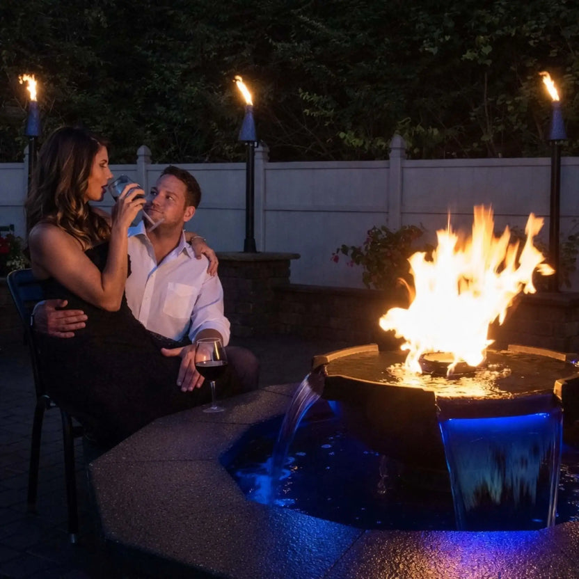 Romantic backyard evening scene featuring HPC Fire Hearth Products H2O n fire fire water feature, where a couple relaxes beside a glowing circular fire and water bowl. The fire burns brightly above cascading water illuminated with blue LED lighting, creating a luxury outdoor ambiance with tiki torches and patio seating.