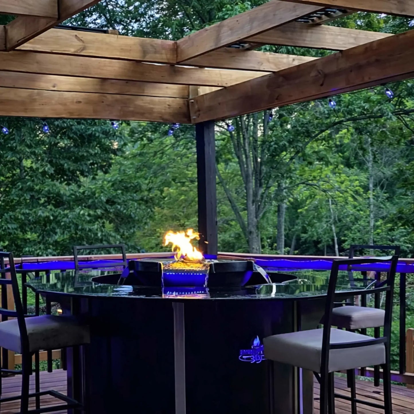 Outdoor deck setup featuring HPC Fire Hearth Products H2O n fire fire water feature built into a circular bar-height table under a wooden pergola. Blue LED lighting accents the water while flames rise at the center, creating a relaxing backyard entertainment space surrounded by trees.