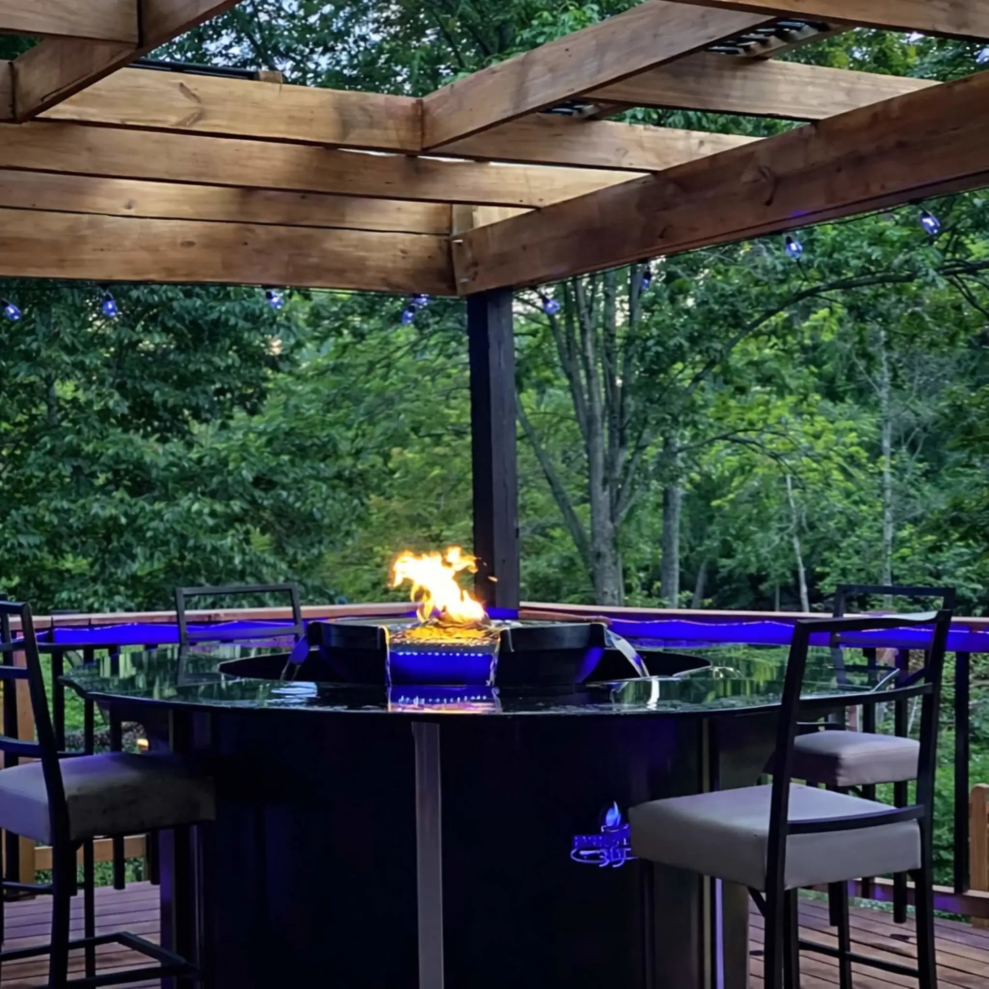Outdoor deck setup featuring HPC Fire Hearth Products H2O n fire fire water feature built into a circular bar-height table under a wooden pergola. Blue LED lighting accents the water while flames rise at the center, creating a relaxing backyard entertainment space surrounded by trees.