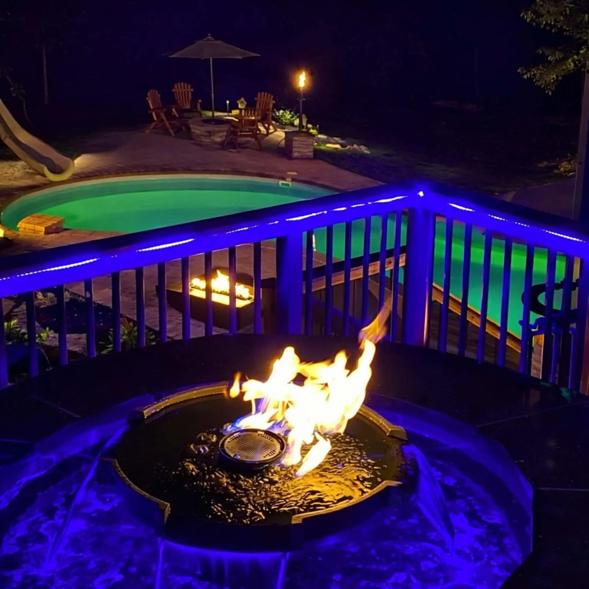 Nighttime backyard deck view featuring HPC Fire Hearth Products Controls H2O n fire fire water feature glowing with blue LED lighting. Flames reflect off the water surface while a nearby pool and patio seating create a warm, inviting outdoor living environment.
