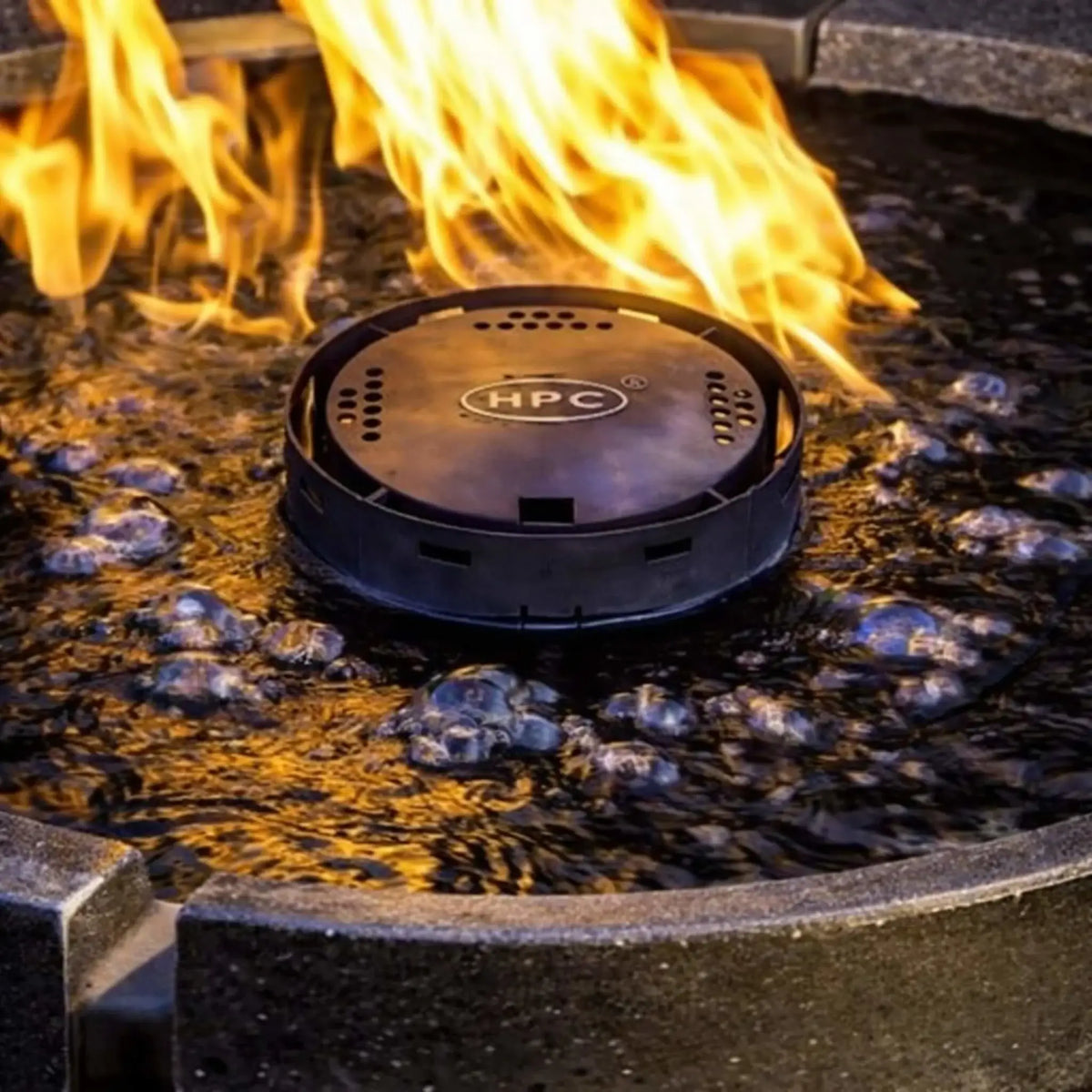Detailed close-up of HPC Fire Hearth Products H2O n fire fire water feature burner cap with visible HPC branding, surrounded by bubbling water and bright flames. The image highlights the precision burner design and dynamic fire-on-water effect.