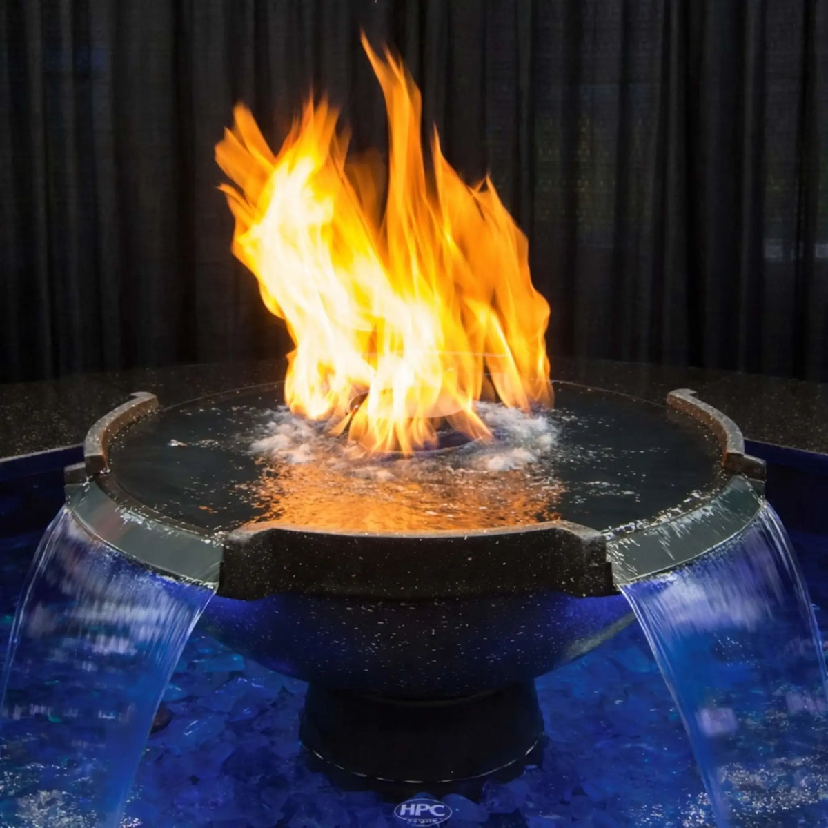 Close-up of HPC Fire Hearth Products Controls H2O n fire fire water feature showcasing vibrant flames rising from a central burner surrounded by flowing water spillways. The textured surface and dual waterfall streams highlight the Vesta Award winning fire and water combination design.
