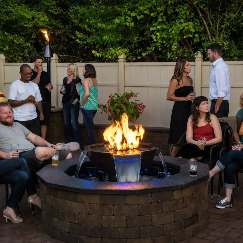 Social backyard party scene centered around HPC Fire Hearth Products Controls H2O n fire fire water feature integrated into a circular stone fire pit table. Flames rise from the center while water cascades into the surrounding basin, with multiple guests relaxing and chatting in a cozy patio environment.
