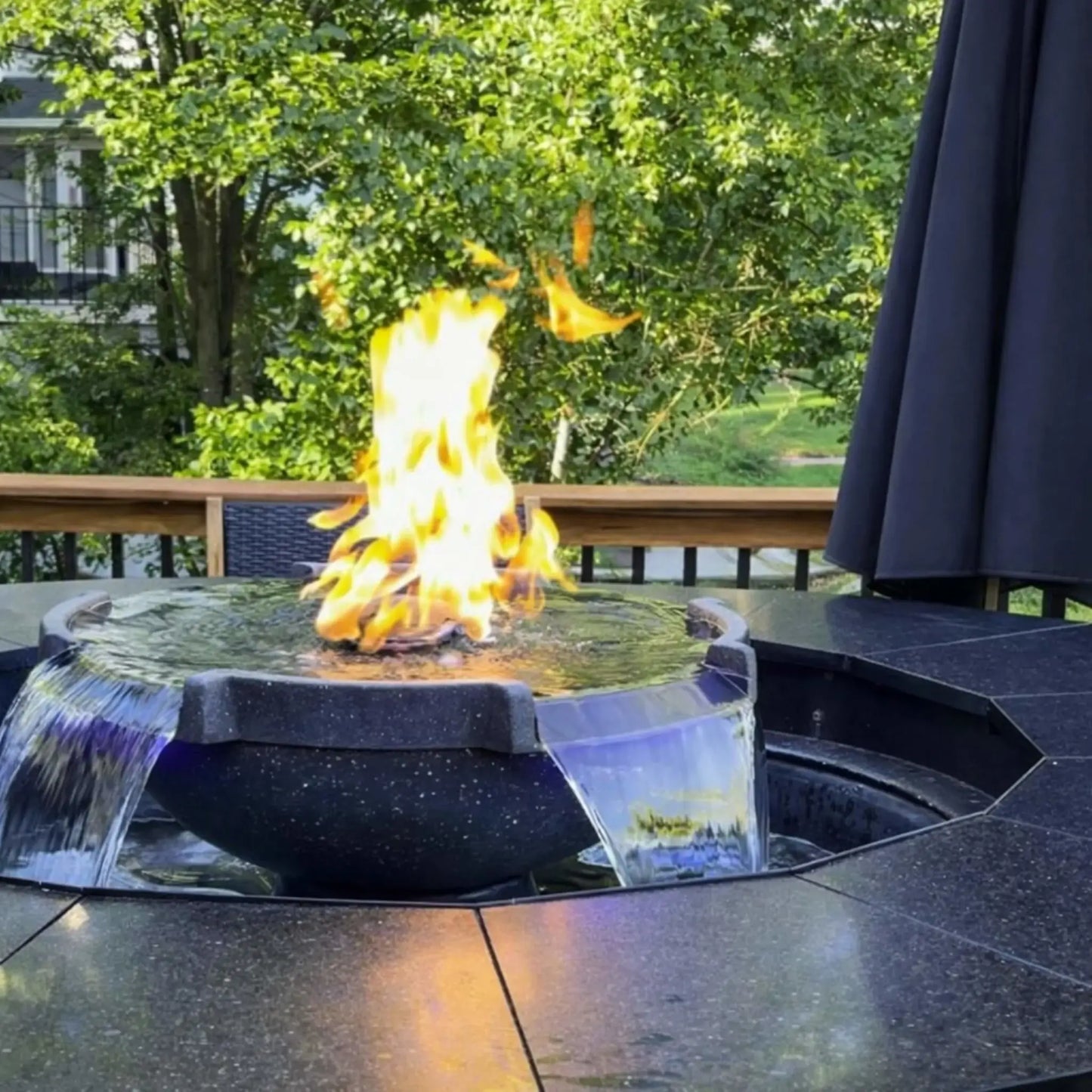 Daytime backyard deck installation of HPC Fire Hearth Products Controls H2O n fire fire water feature showing a circular unit with active flame and dual water spillways. The fire sits above rippling water while greenery and patio furniture create a serene residential outdoor setting.