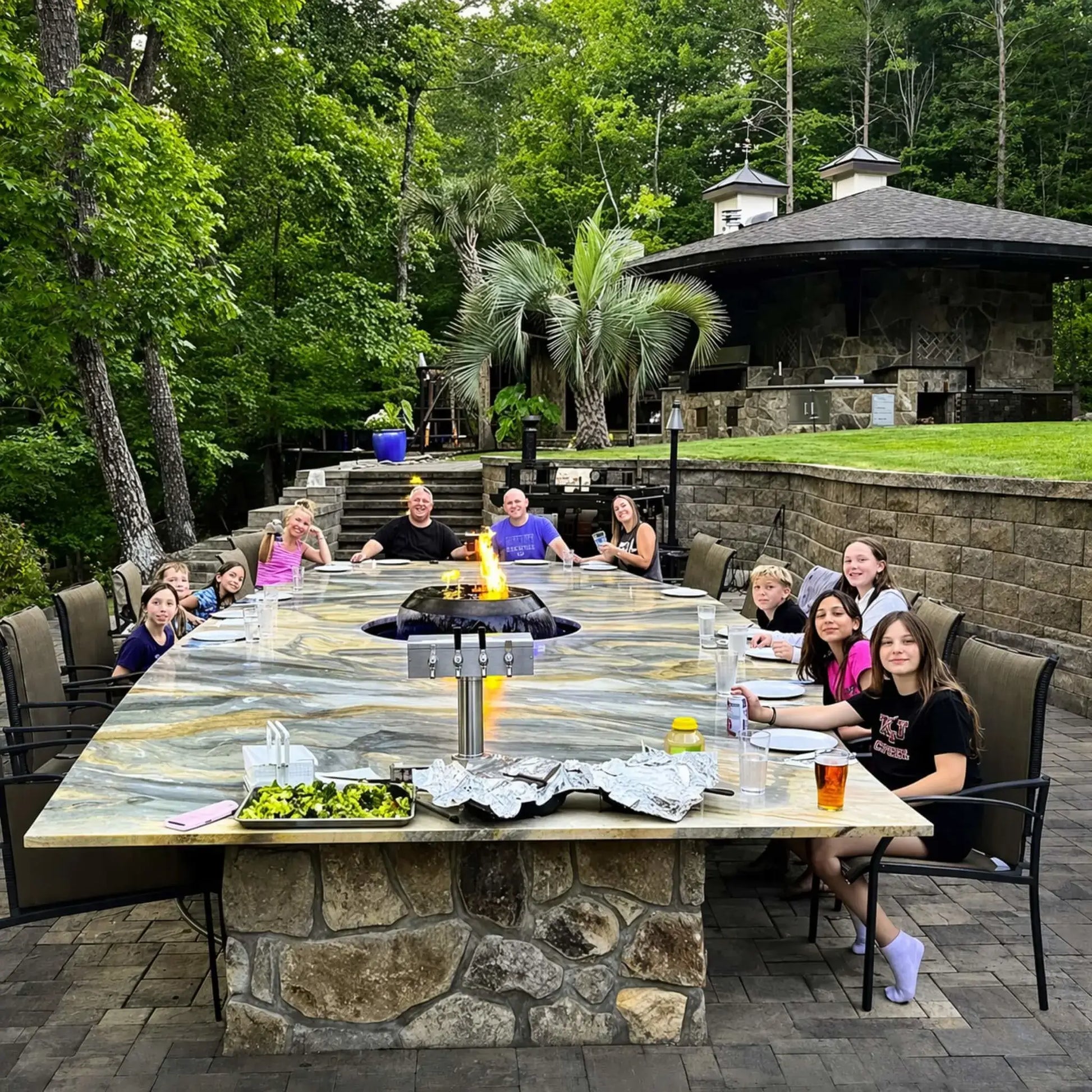 Large outdoor dining area featuring HPC Fire Hearth Products H2O n fire fire water feature integrated into a long stone table. Family and friends gather around the centerpiece where flames rise above water, combining fire and water elements for a unique luxury outdoor dining experience.