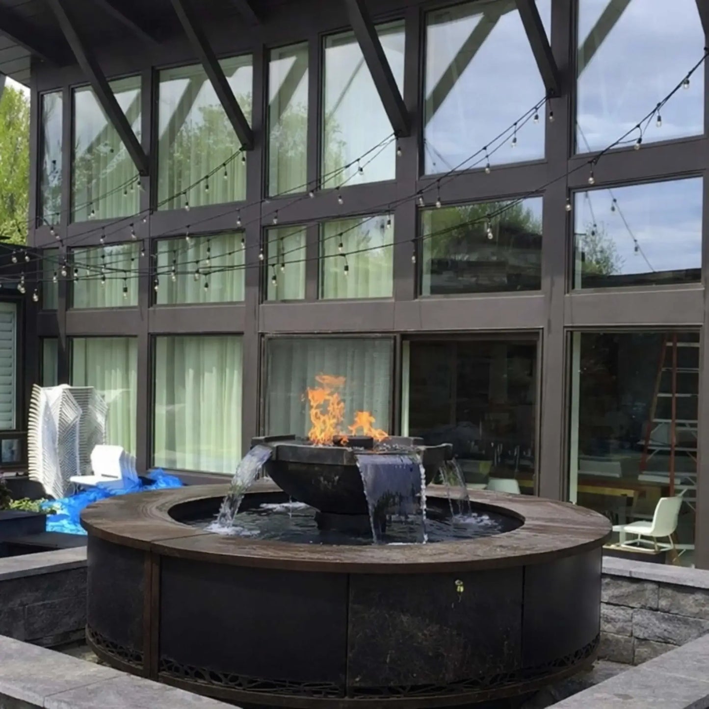 Contemporary courtyard installation of HPC Fire Hearth Products Controls H2O n fire fire water feature set within a circular metal enclosure. Flames burn above cascading water streams, complemented by modern architecture, large windows, and string lighting for an upscale outdoor design.
