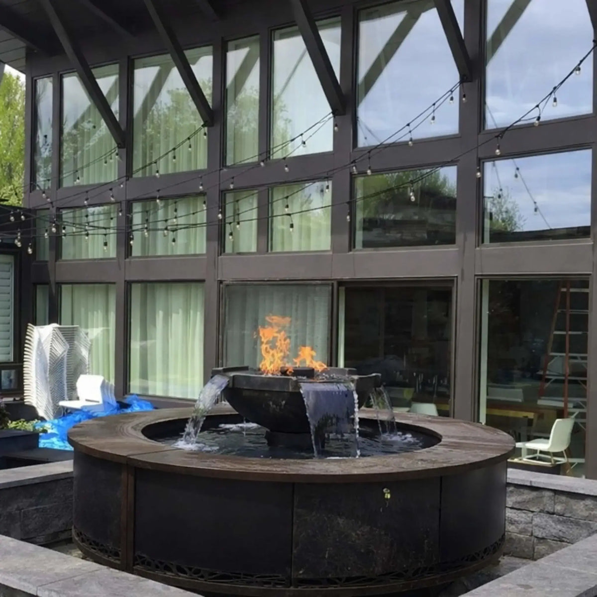 Contemporary courtyard installation of HPC Fire Hearth Products Controls H2O n fire fire water feature set within a circular metal enclosure. Flames burn above cascading water streams, complemented by modern architecture, large windows, and string lighting for an upscale outdoor design.