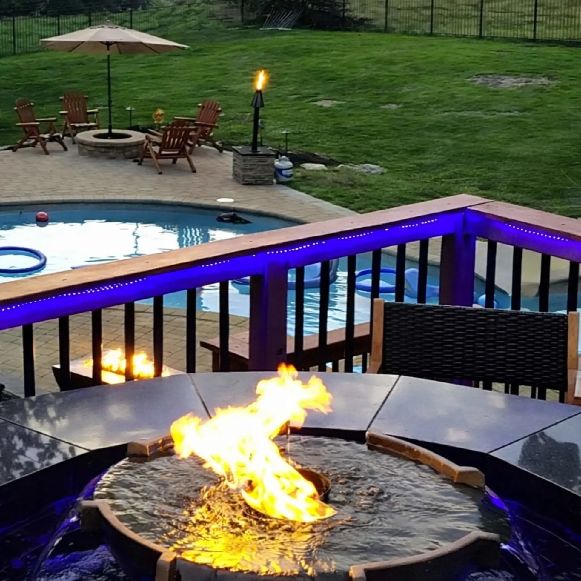 Backyard deck overlooking a pool with HPC Fire Hearth Products H2O n fire fire water feature as the focal point. Flames rise from the center while water flows over the edges, enhanced by blue LED lighting and surrounding patio seating for a relaxing evening outdoor environment.