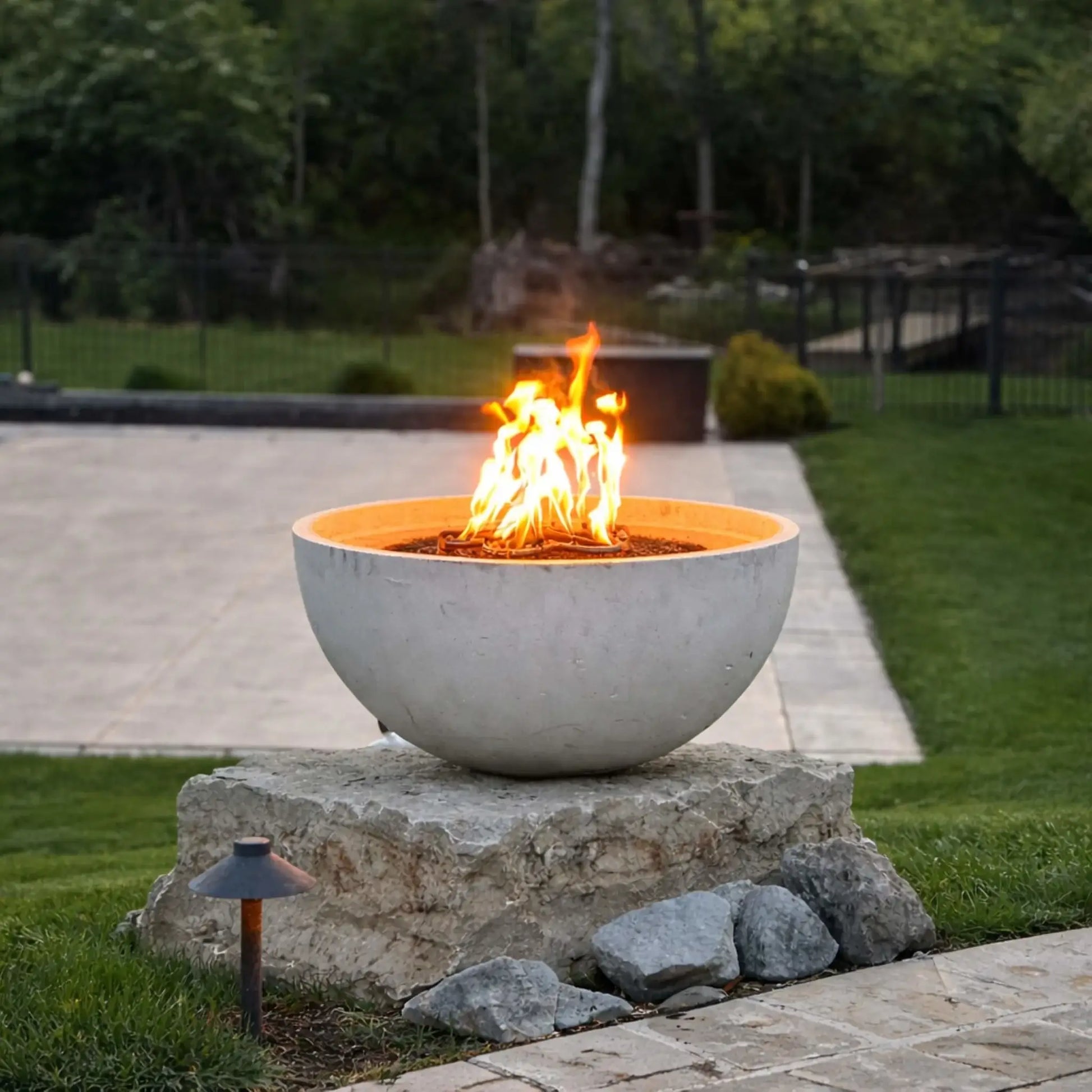 Contemporary concrete fire bowl mounted on a stone pedestal using High Capacity Match Lit Ignition Round Flat Pan burner, displaying steady flames in a manicured backyard with lawn, patio, and subtle landscape lighting.