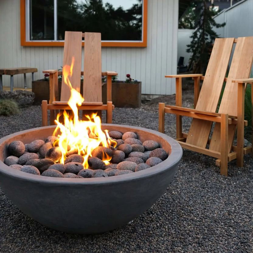 Contemporary backyard fire pit with round bowl design and match lit ignition burner, surrounded by wooden chairs and gravel landscaping, showcasing lively flames and natural stone fire media in a relaxed outdoor seating area.
