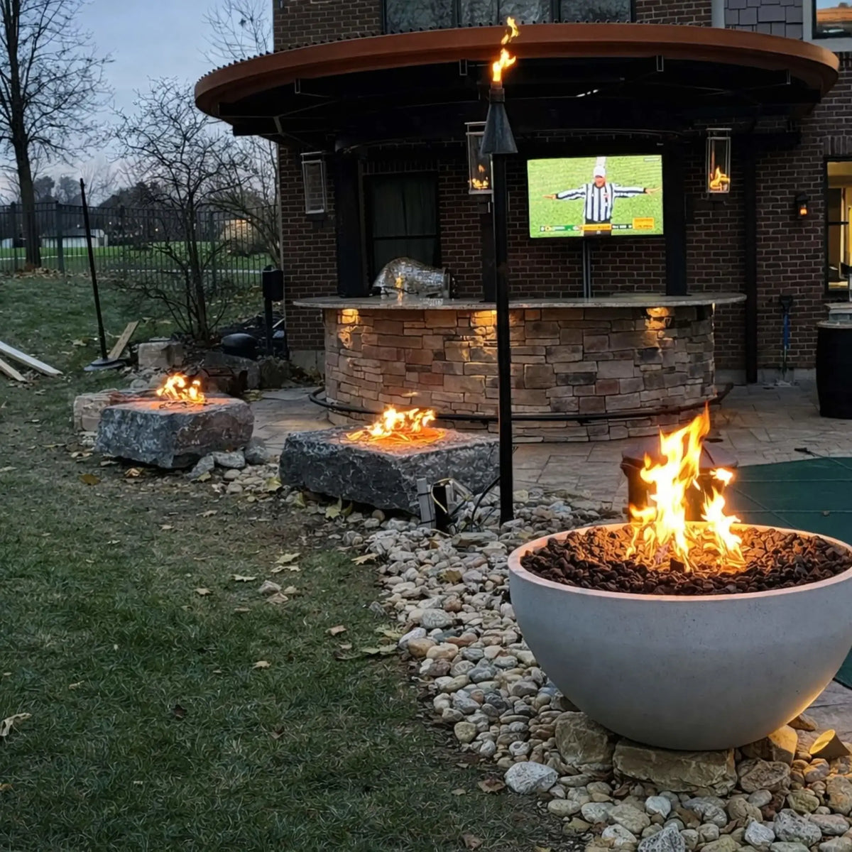 Outdoor backyard entertainment area featuring multiple fire features using High Capacity Match Lit Ignition Round Flat Pan burner systems, including a concrete fire bowl and stone fire pits leading toward a covered bar with TV, highlighting warm flames and premium HPC Fire Hearth Products Controls setup.