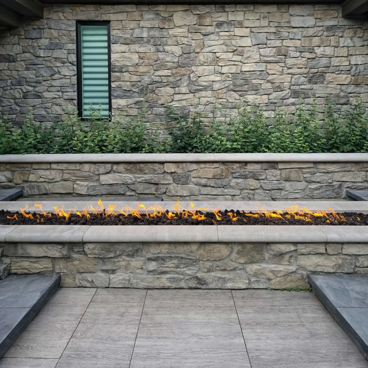 Linear outdoor gas fire feature built into a stone retaining wall, filled with black lava rock and producing a clean row of flames, set against a textured natural stone backdrop with a narrow vertical window and surrounding greenery for a modern landscape design.