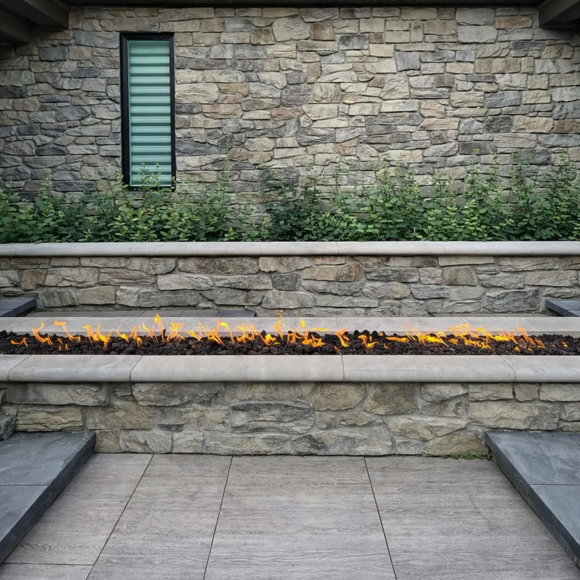 Linear outdoor gas fire feature built into a stone retaining wall, filled with black lava rock and producing a clean row of flames, set against a textured natural stone backdrop with a narrow vertical window and surrounding greenery for a modern landscape design.