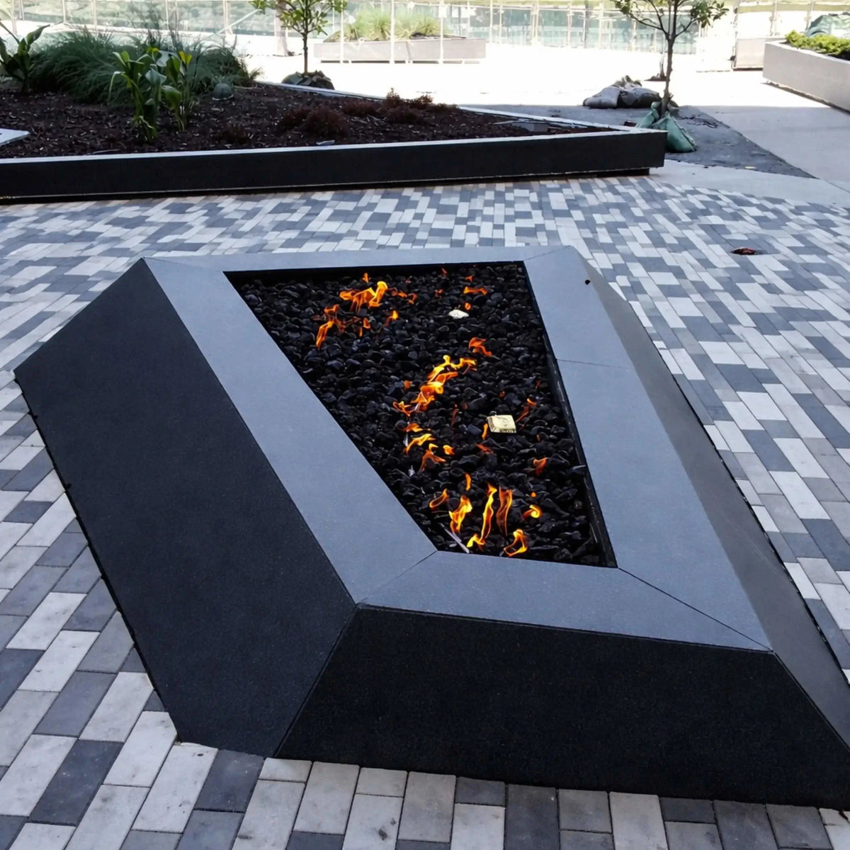 Modern triangular gas fire pit with a matte black metal frame, filled with black lava rock and scattered flames, set on a patterned gray paver plaza with raised planter beds and minimal landscaping in a contemporary outdoor commercial space.