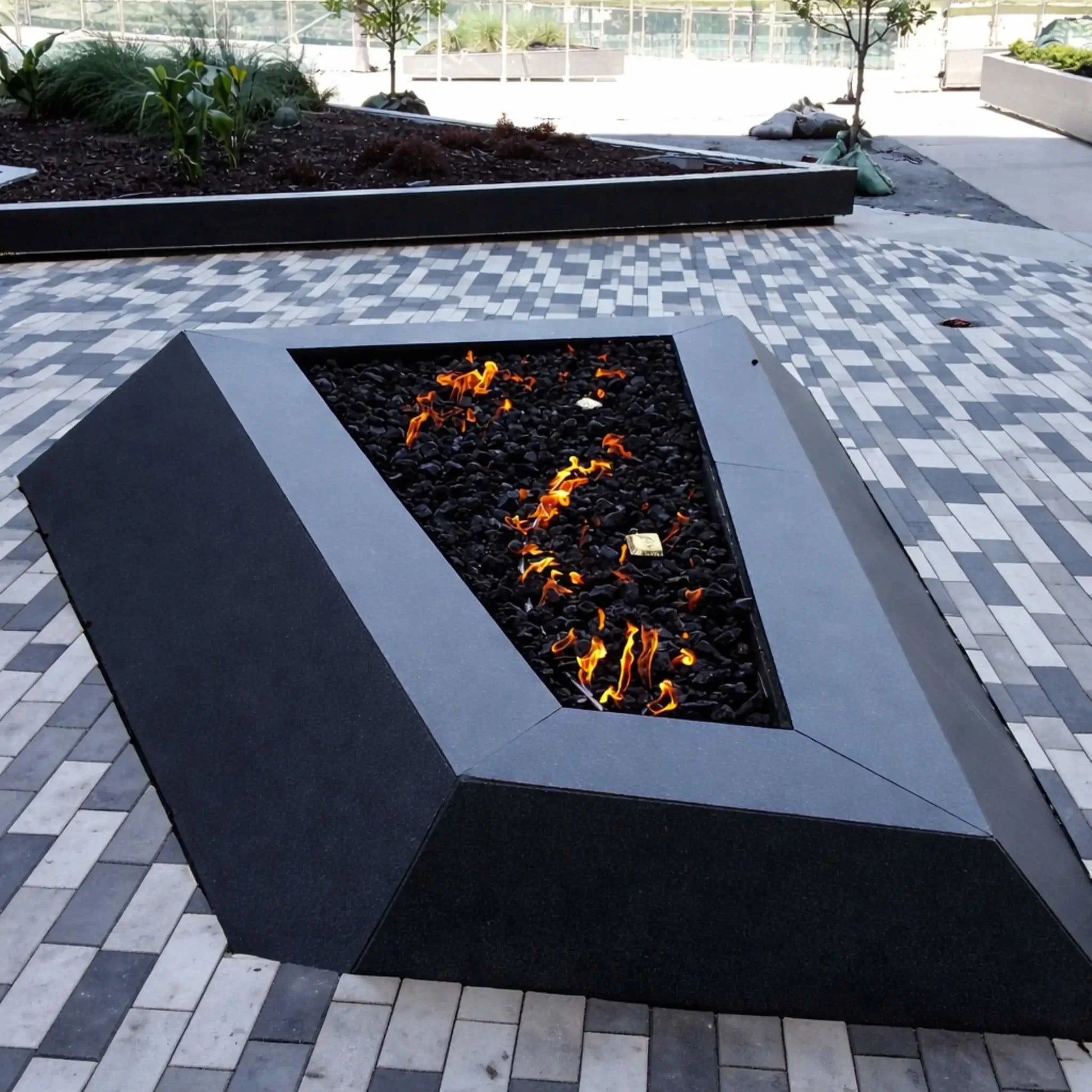 Modern triangular gas fire pit with a matte black metal frame, filled with black lava rock and scattered flames, set on a patterned gray paver plaza with raised planter beds and minimal landscaping in a contemporary outdoor commercial space.