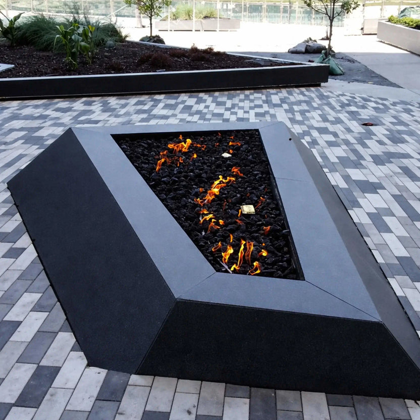 Modern triangular gas fire pit with a matte black metal frame, filled with black lava rock and scattered flames, set on a patterned gray paver plaza with raised planter beds and minimal landscaping in a contemporary outdoor commercial space.