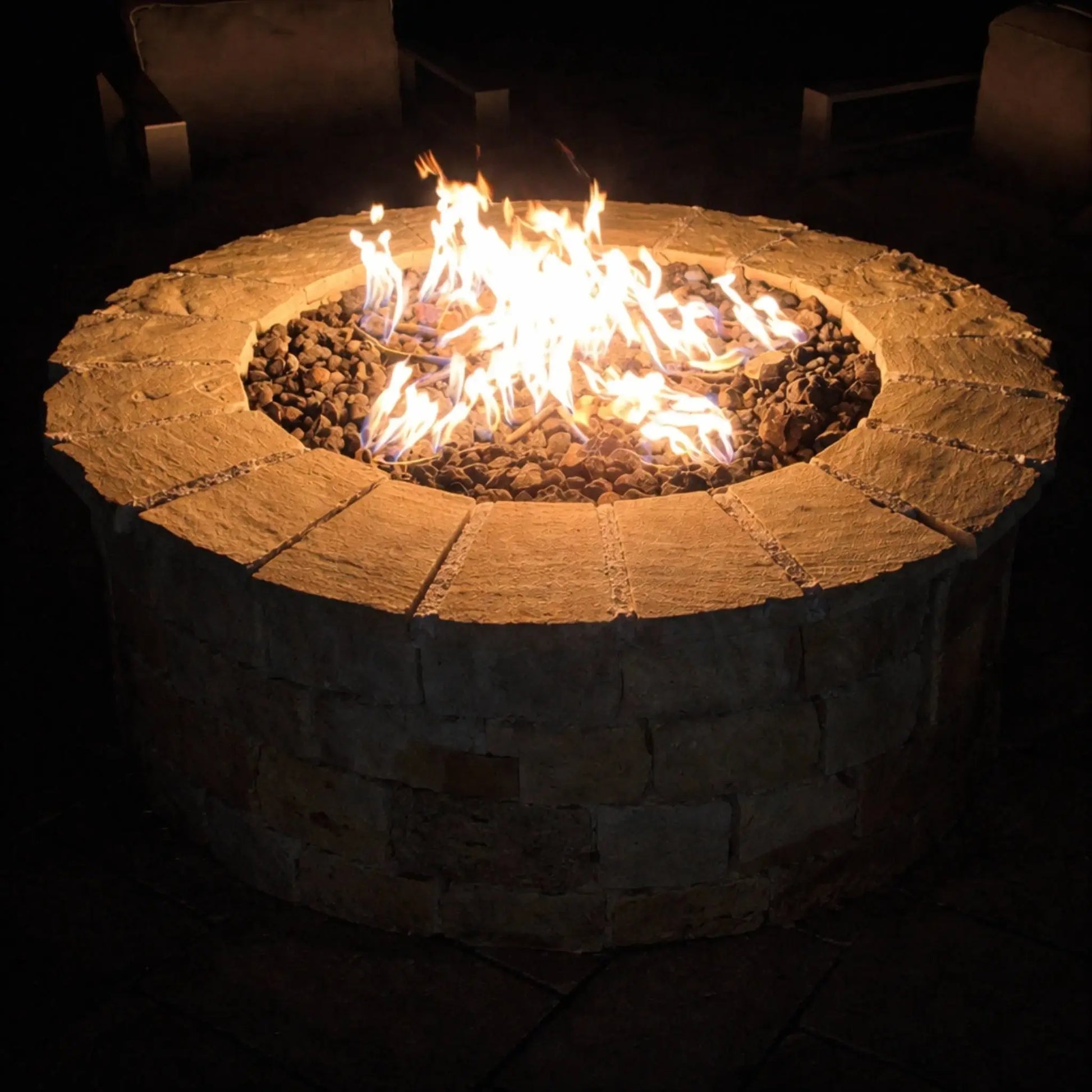 Round stone fire pit glowing at night, filled with lava rock and bright, natural gas flames, surrounded by cushioned outdoor seating in a dark, cozy patio setting that highlights the warmth and ambiance of the fire feature.