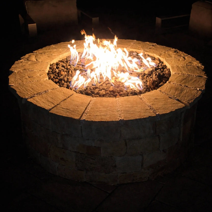 Round stone fire pit glowing at night, filled with lava rock and bright, natural gas flames, surrounded by cushioned outdoor seating in a dark, cozy patio setting that highlights the warmth and ambiance of the fire feature.