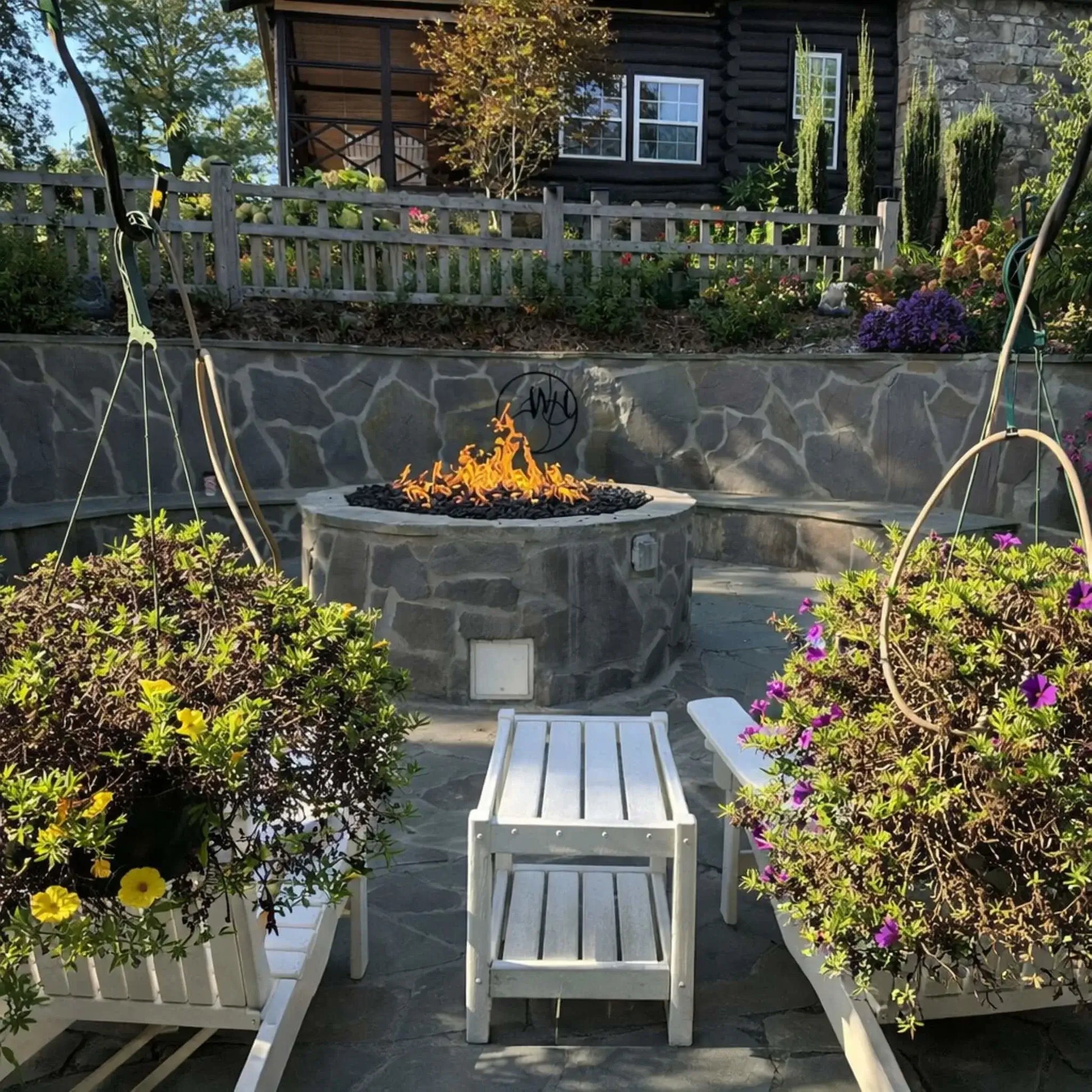 Charming backyard garden patio featuring a circular stone fire pit with lava rock and steady flames, framed by hanging flower baskets, colorful blooms, and a rustic wooden fence in front of a cabin-style home.