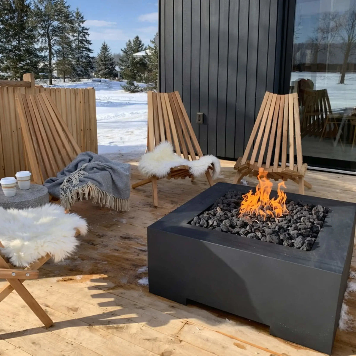 Contemporary square gas fire pit table with a dark finish and lava rock burner, surrounded by wooden lounge chairs with white fur throws on a snow-covered patio, creating a warm outdoor winter seating area.