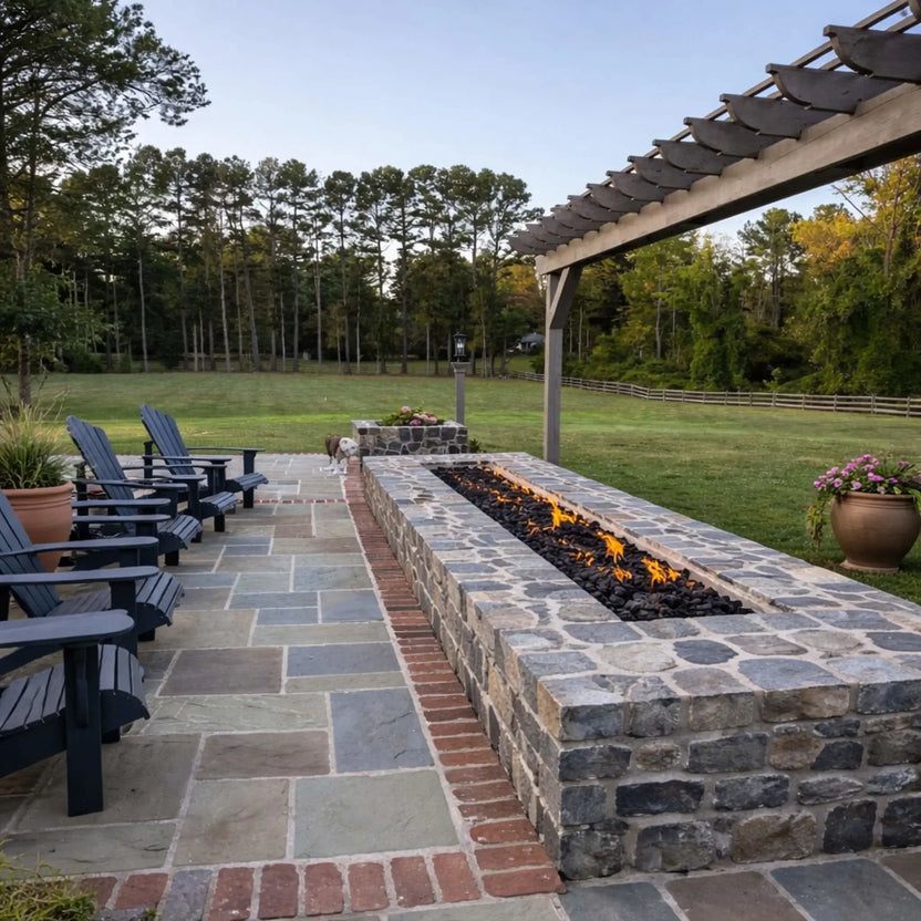 Long linear gas fire feature built into a stone structure beneath a wooden pergola, filled with lava rock and producing a clean line of flames, overlooking an open grassy landscape with Adirondack chairs nearby.