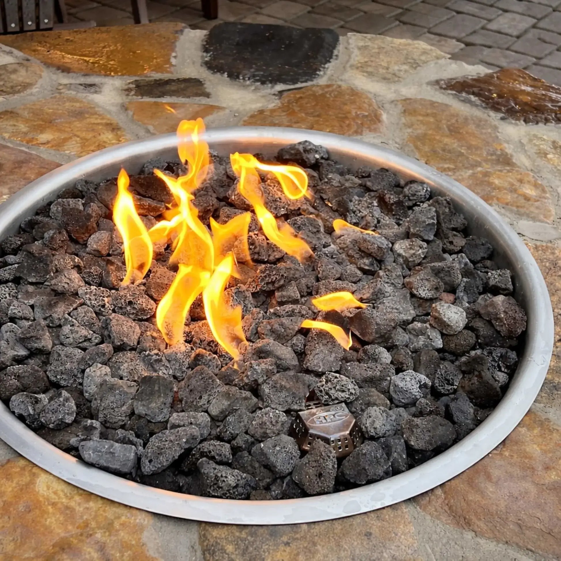 Close-up of a circular gas burner pan filled with black lava rock and active flames, set into a natural stone tabletop, highlighting the burner components and texture of the lava rock media.