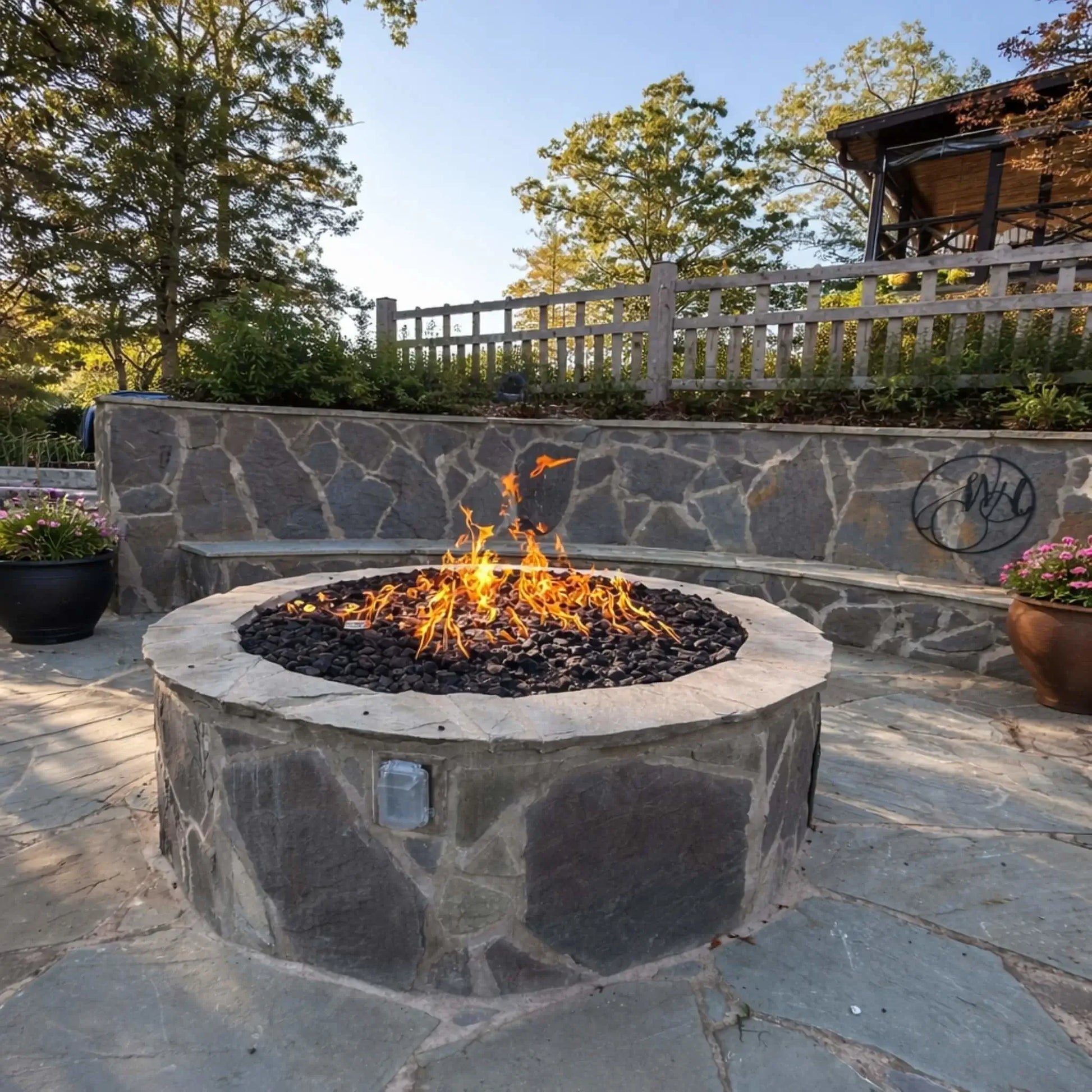 Custom circular stone fire pit with black lava rock and vibrant flames, positioned in a landscaped patio with curved stone seating walls, planters, and a decorative fence in a peaceful backyard setting.