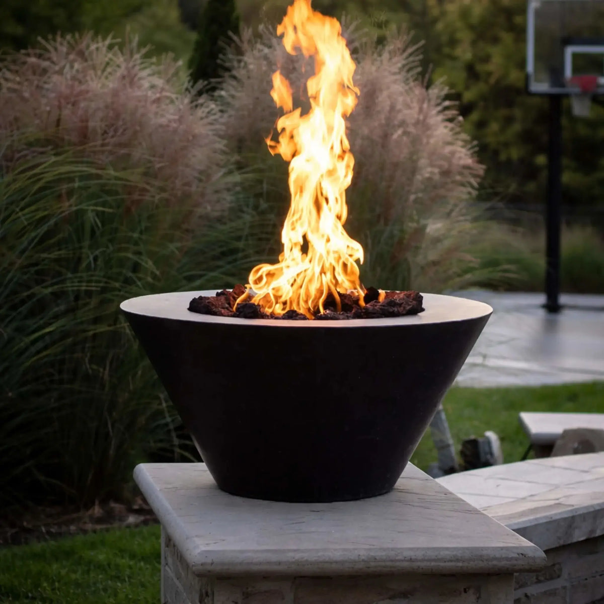 Modern black metal fire bowl with a tall vertical flame rising from lava rock, mounted on a stone pillar in a landscaped yard with ornamental grasses and a basketball hoop in the background.