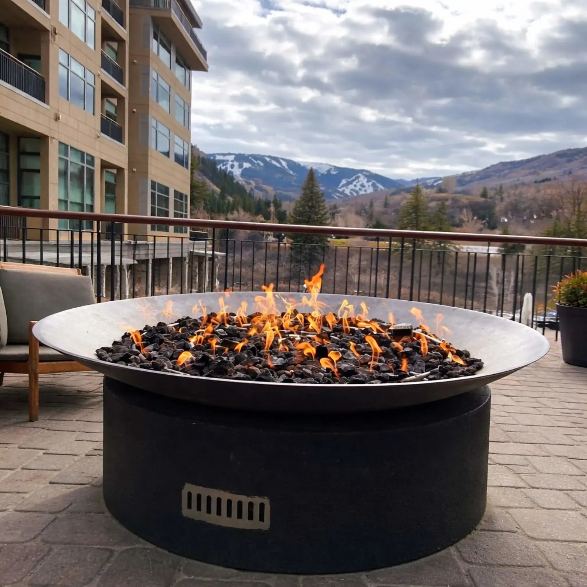 Large round metal gas fire bowl filled with black lava rock and lively flames, positioned on a paved patio balcony overlooking a mountain landscape with snow-dusted peaks, modern buildings, and a cloudy sky in the background.