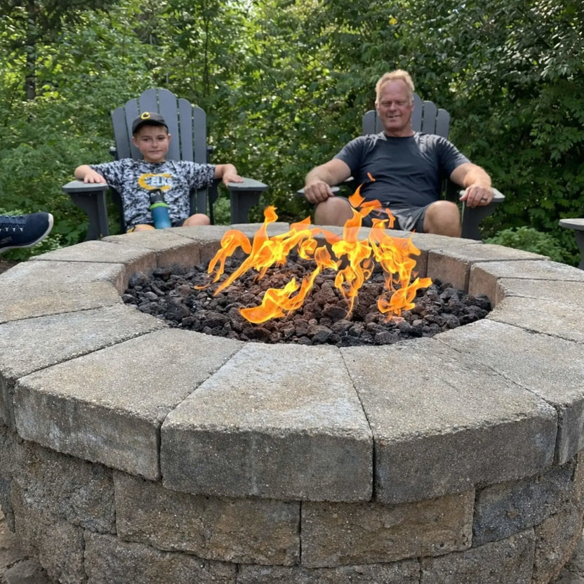 Circular stone fire pit filled with lava rock and bright flames, surrounded by Adirondack chairs with a man and child relaxing beside it in a wooded backyard setting, creating a warm and inviting family gathering space.