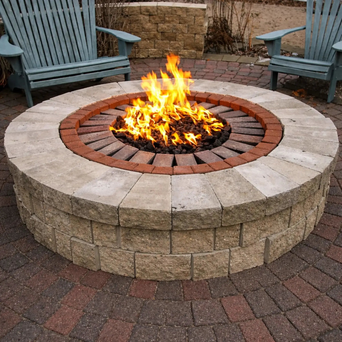 Round stone fire pit with a decorative brick ring and lava rock center, producing strong flames, set on a circular paver patio with outdoor chairs arranged around it in a landscaped backyard.