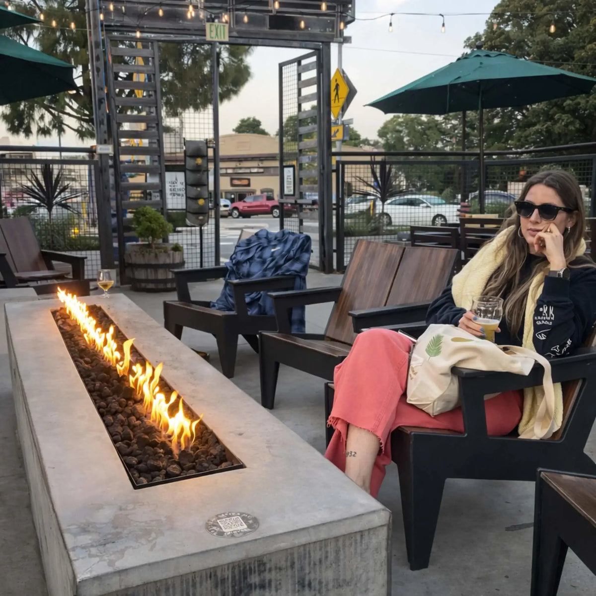 Outdoor patio lounge area featuring a long linear gas fire table filled with lava rock and steady flames, with a woman sitting nearby holding a drink, surrounded by modern seating, string lights, and urban street views.