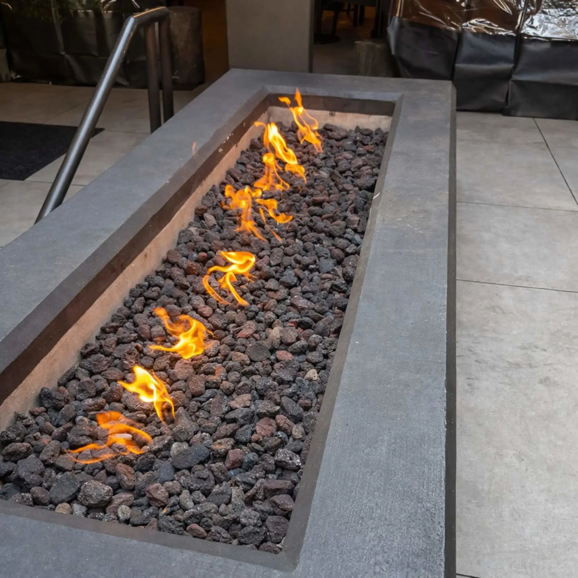 Close-up of a rectangular gas fire feature filled with black lava rock, showing active flames along a stainless steel burner tube, highlighting ignition components and media texture within a concrete fire pit enclosure.