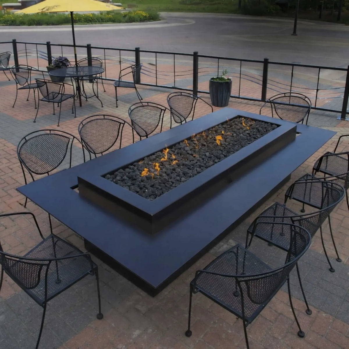 Modern outdoor living space with multiple fire features, including stone fire pits and a fire bowl, arranged along a landscaped backyard with decorative rock beds, leading toward a covered outdoor bar area with a mounted TV.
